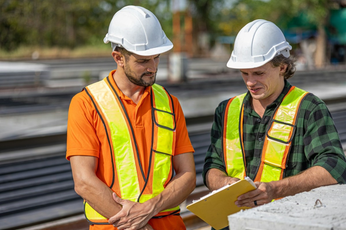 Two construction workers in hard hats and hi-vis vests reviewing plans on a clipboard on site